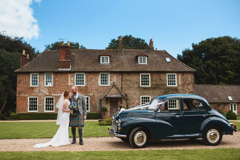 A just married couple enjoy a moment next to a vintage Morris Minor outside Solton Manor, Dover, Kent, England, UK